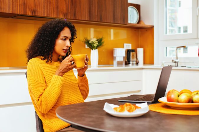 Woman in colorful kitchen sipping from mug in front of her laptop