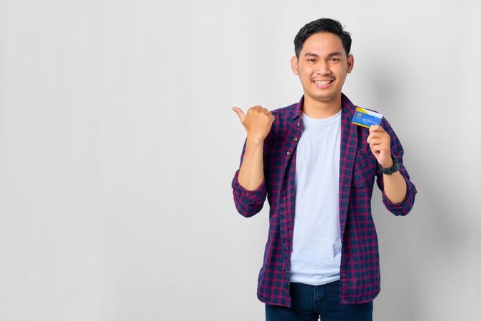 Asian male in grey studio with pointing thumb to the side with credit card