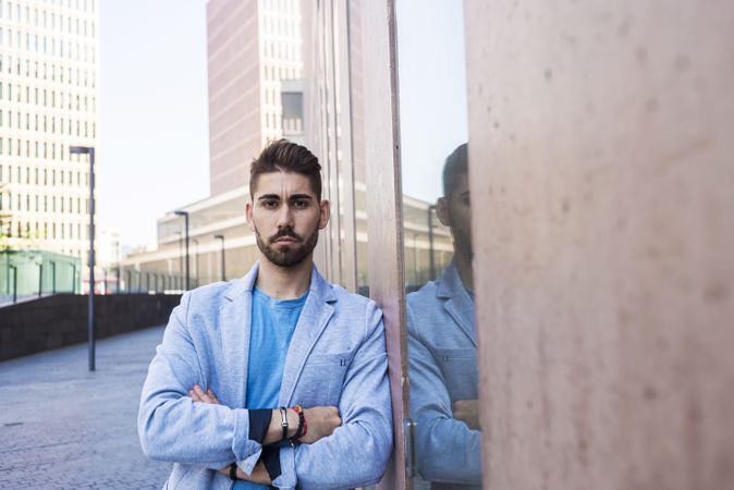 Young man in blazer with arms crossed leaning on a building wall outside looking at camera