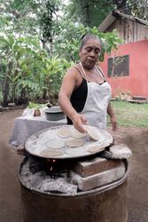 A Hispanic woman is cooking traditional corn tortillas over an outdoor stove