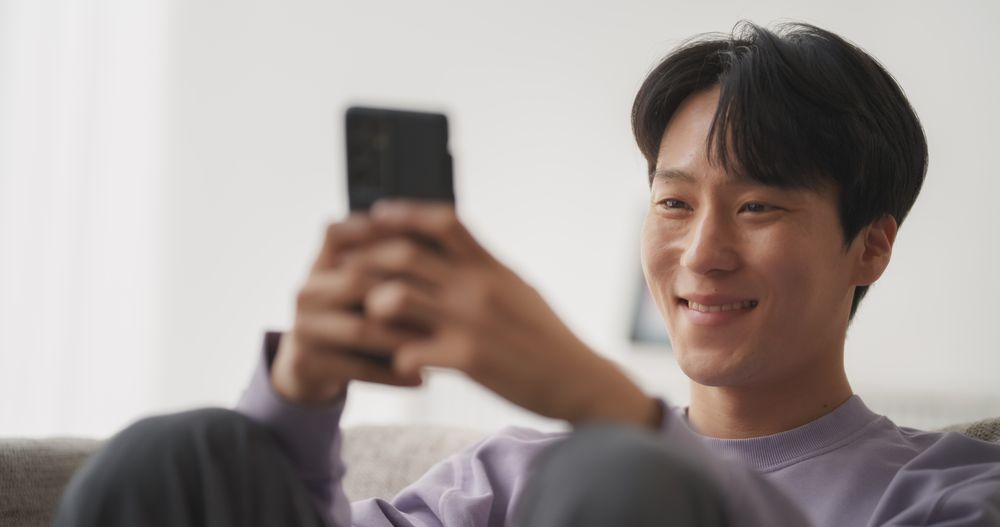 Young Asian Male Browsing Internet, Using Smartphone at Home