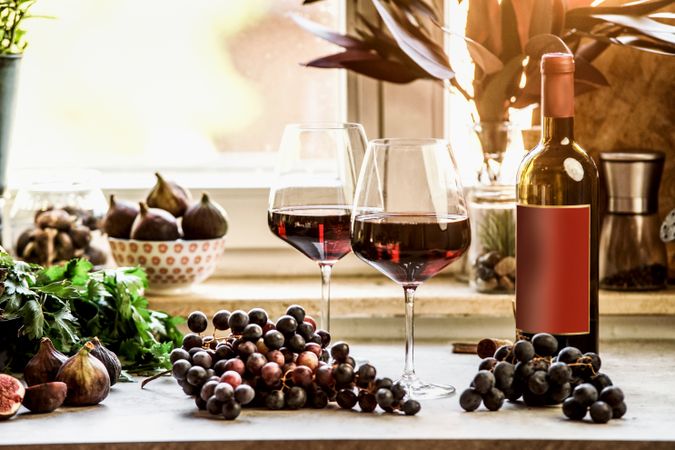 Red wineglasses with grapes and wine bottle at kitchen table at window background. Front view. Copy space.