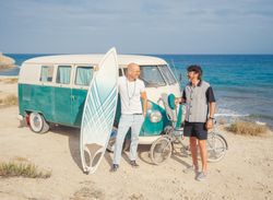 Men with surfboard and bicycle standing near car on dusty seaside