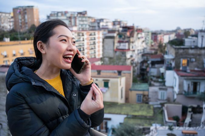 Joyful Asian female tourist in warm wear calling on smartphone at big old city