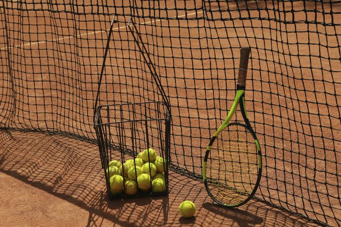 Basket with tennis balls and racket against net on clay court
