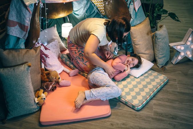 Top view of young daughter laughing while mother tickling in a diy tent camping at home