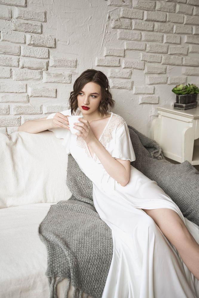 Pensive adult woman looking away and dreaming while sitting on sofa and having hot drink in light modern living room