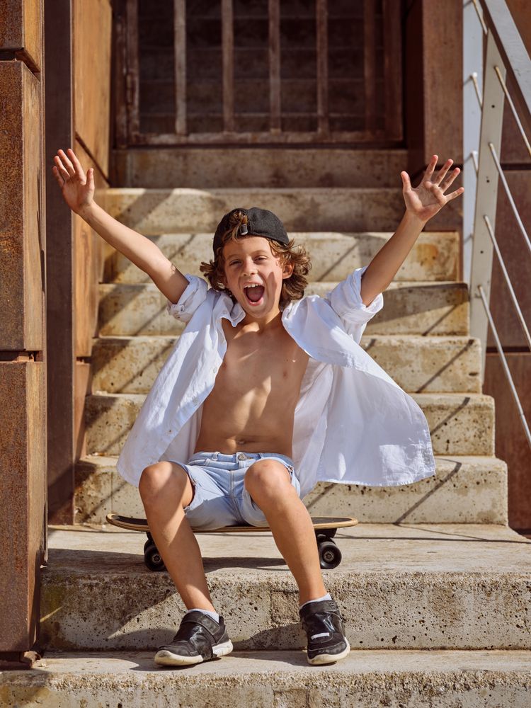 Cheerful boy in summer outfit with raised hands sitting on stair