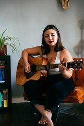Young woman sitting barefoot in orange chair holding guitar with eyes closed
