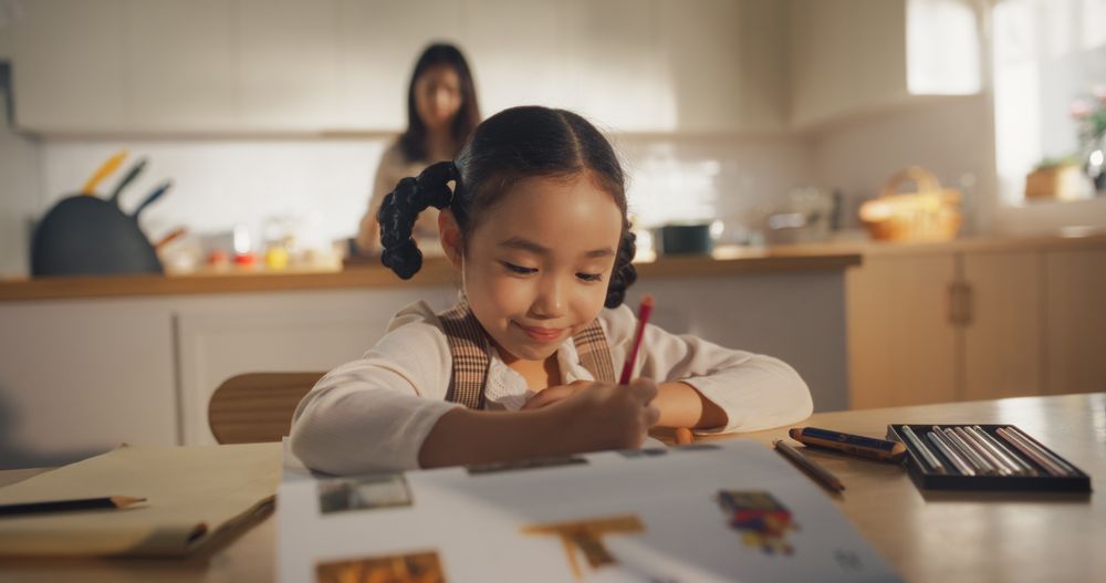 Portrait of a Korean Female Child Sitting at a Kitchen Table and Drawing While her Mother is Preparing Breakfast.