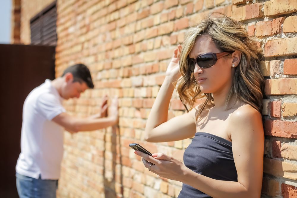 Angry woman looking phone and man posing on wall