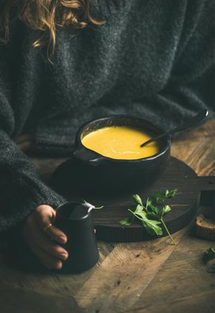Blonde woman with warm yellow soup in dark bowl holding cream