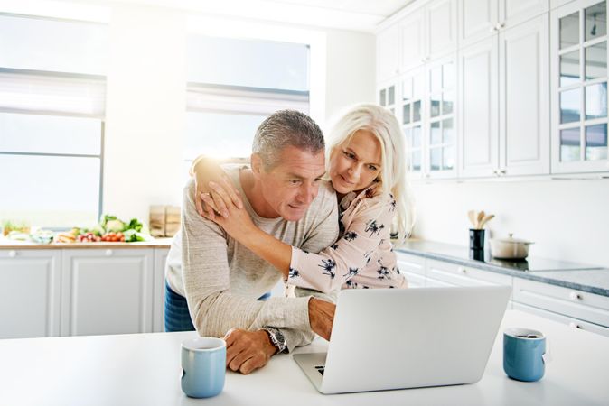 Grey haired couple embracing in the the kitchen with a laptop and cups of tea
