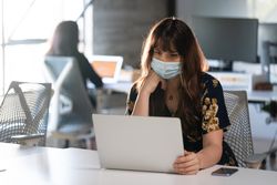 Transgender woman wearing a face mask working at her desk in an open plan office