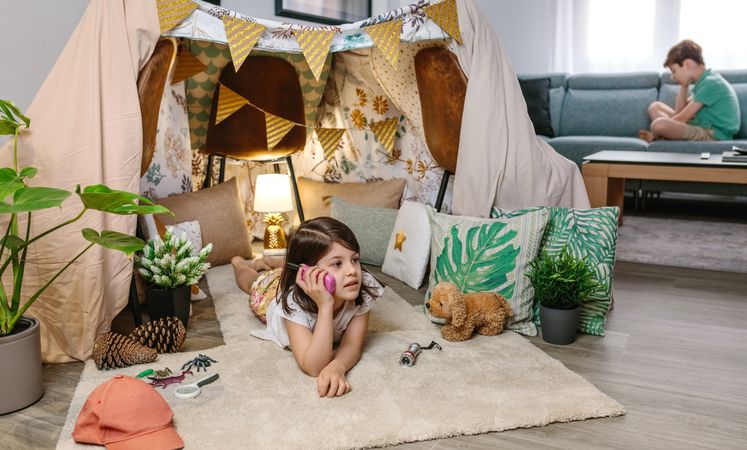 Little girl holding walkie-talkie lying over rug in shelter tent while talk with boy sitting on sofa