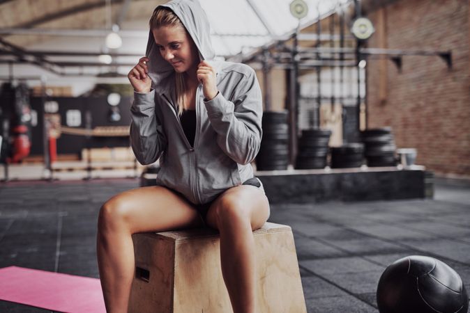Woman sitting on box in gym grasping her hood while smiling