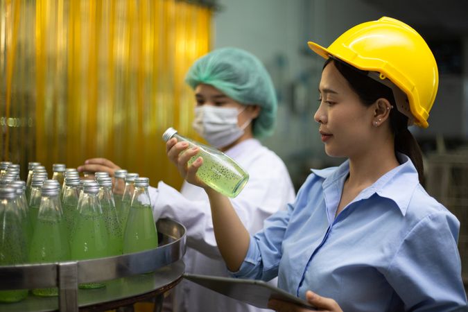 Asian female manager and worker holgin digital tablet checking information and quality control for juice drink product on bottling factory machinery.