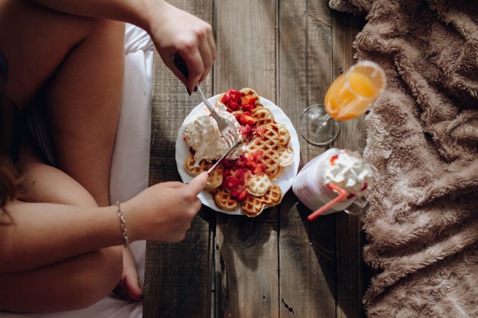 Young woman eating appetizing waffles and tasty drinks on tray on bed