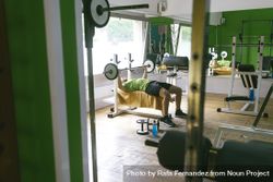 Looking through gym equipment at man in green t-shirt lifting heavy bar exercising arms and chest 0WW7O0
