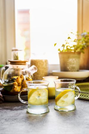 Healthy beverage with lemon and ginger in glass tea cups at kitchen table with mugs, candle and herbs at window background with natural light. Warming hot drinks with citrus fruit. Front view.