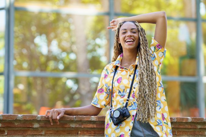 Smiling female in bold patterned shirt leaning on brick wall with camera and arm above head