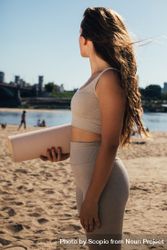 Side view of woman holding a rolled yoga mat standing on beach 43EQZ0