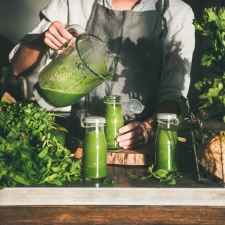 Woman pouring green smoothie into jars in kitchen, square crop