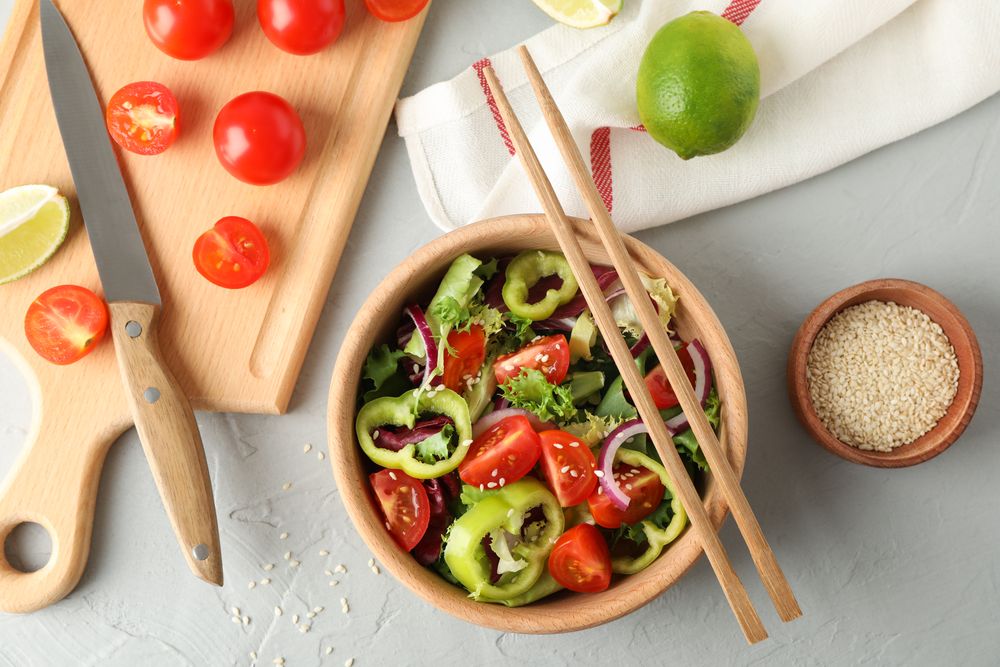 Fresh salad with chopsticks and kitchen accessories on grey background, top view