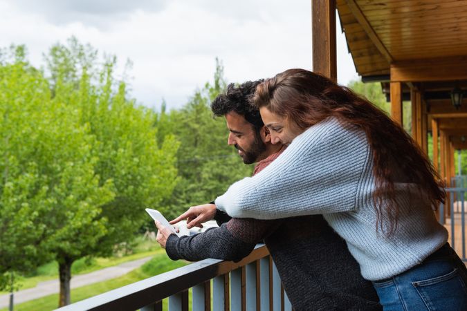 Cheerful traveling couple embracing and using smartphone on terrace of house