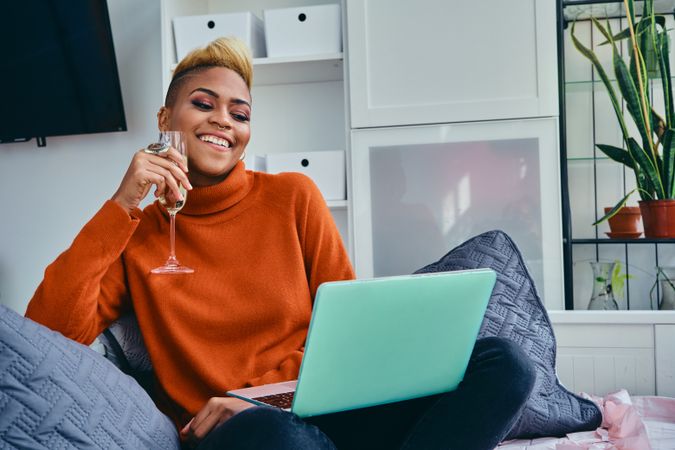 Happy Black woman sipping champagne while talking on a laptop in a bright studio loft