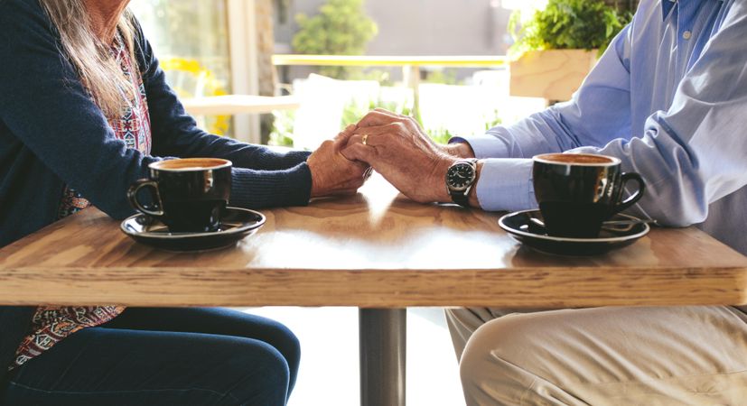 Cropped shot of older couple sitting at a coffee shop table holding hands with two coffee cups