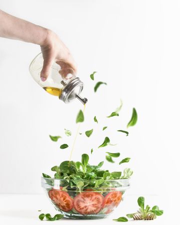Woman hand pouring olive oil over green salad in glass bowl with tomatoes and flying lettuce leaves
