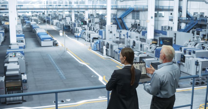Male Engineer and Female Scientist Standing with Their Back, Using Laptop Computer and Talking in a Factory Facility with Equipment Producing Modern Technical Devices for Different Industries