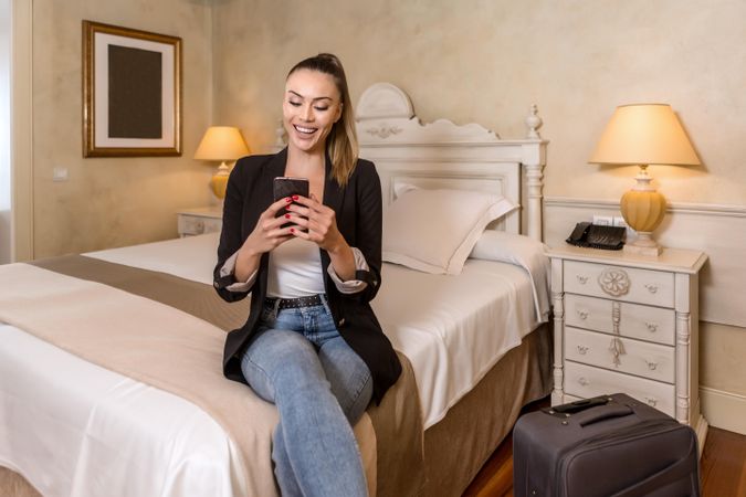 Smiling woman in dark blazer using smartphone and sitting on bed beside luggage in hotel room