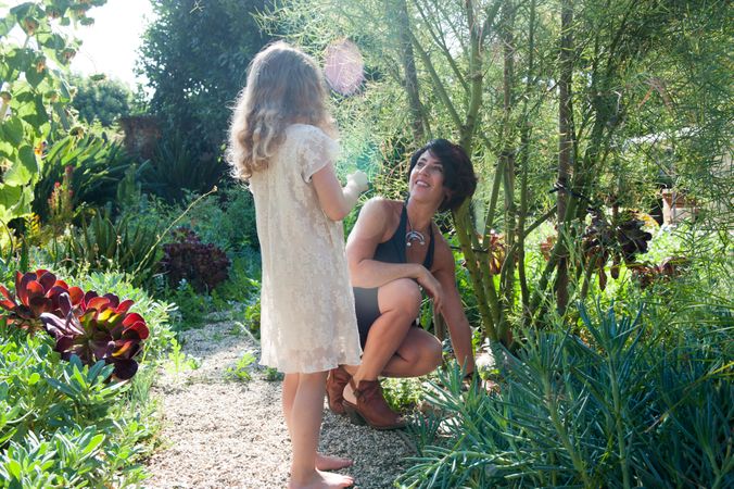 Mother sitting on rock path smiling at young daughter in white dress