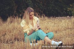 Blonde woman using smartphone sitting on brown grass 0WLXx5