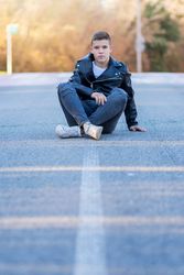 Teenage male wearing a leather jacket sitting crossed legged on concrete looking at camera