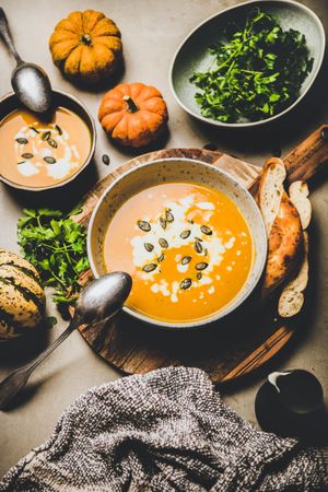 Two bowls of pumpkin soup with garnishes, bread, cream, squash, on concrete surface