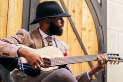 Stylish Black man in hat and classic outfit playing guitar outdoors