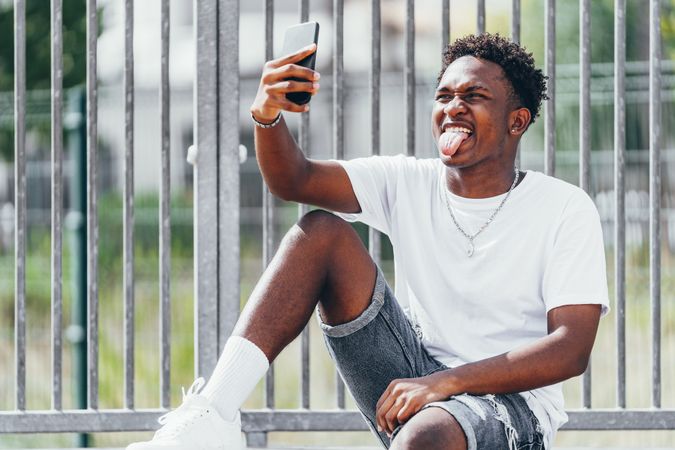 Sportive black men thoughtfully browsing smartphone while sitting on fence