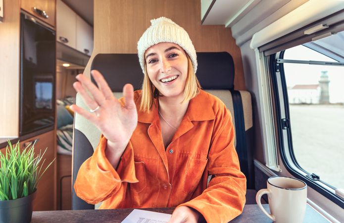 Woman in orange shirt sitting and waving in van with smartphone