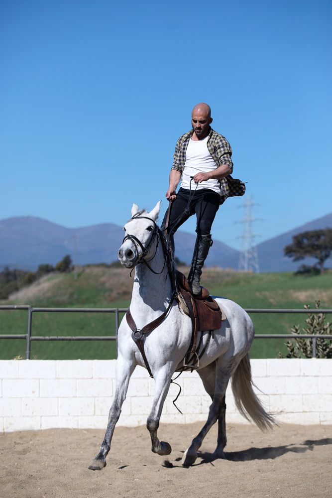 Man in checkered shirt balancing on saddle while riding horse