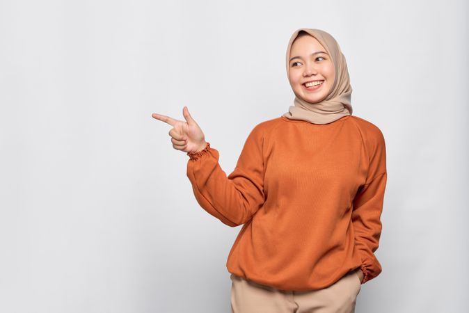 Confident Muslim woman in headscarf and orange sweater pointing finger to blank copy space