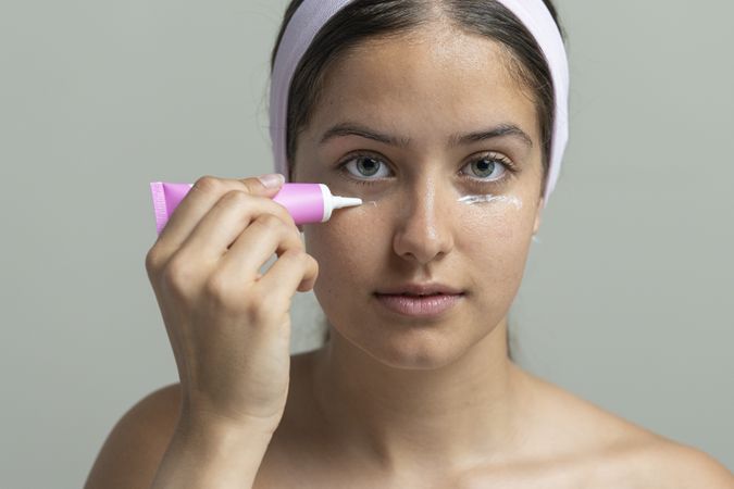 Young woman applying eye cream from a tube, skincare beauty routine on a studio background.