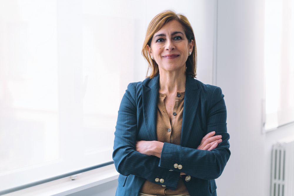 White businesswoman in her 50s, in office attire, standing near window in office, with arms crossed, smiling at camera