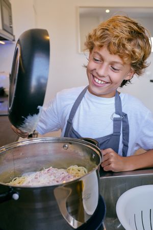 Content boy adding carbonara sauce to cooked spaghetti in kitche