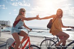 Female friends touching hands and riding bicycles along a seaside promenade on a summer day bEN6G0