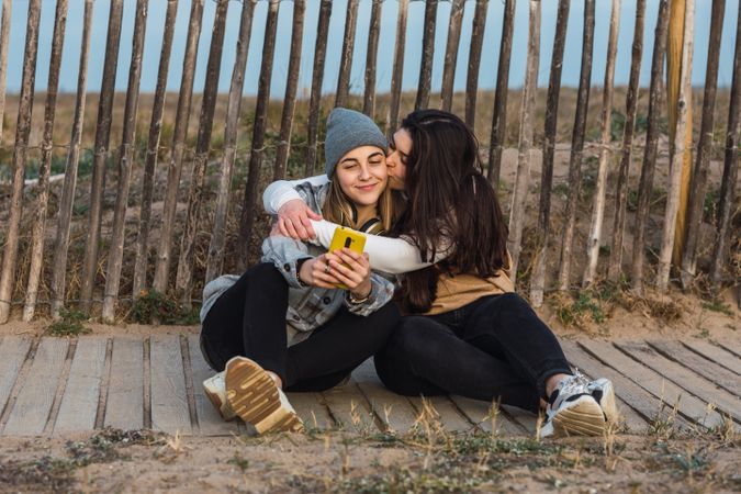 Young women kissing cheek of girlfriend while resting together on boardwalk in nature