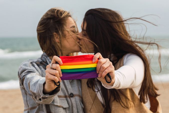 Young lesbians kissing and showing colorful LGBT mask on seashore