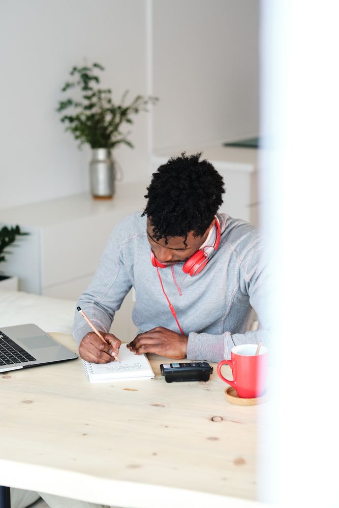 Black student with headphones writing in notebook at home